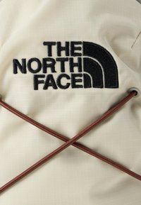 Λευκό ύφασμα με το μαύρο κεντημένο λογότυπο The North Face και δύο καφετί ελαστικά κορδόνια που διασταυρώνονται, περασμένα μέσα από μεταλλικά κουμπώματα.
