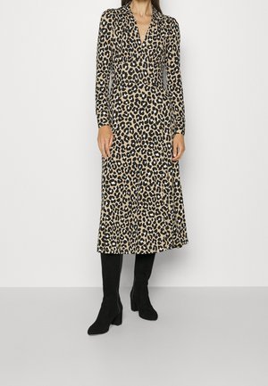 Frau trägt ein langärmliges, knielanges Kleid mit Leopardenmuster, kombiniert mit schwarzen, halbhohen Stiefeln mit Absatz, steht vor einem schlichten weißen Hintergrund.