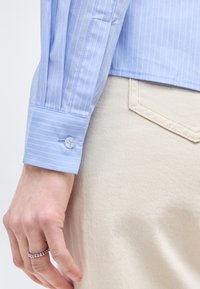 Χέρι με ασημένιο δαχτυλίδι με χάντρες να ξεκουράζεται κοντά στην τσέπη μπεζ παντελονιού, φορώντας μανσέτα μπλε ριγωτού πουκαμίσου με κουμπιά.