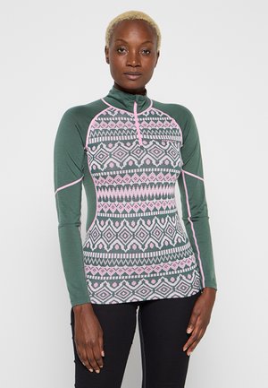 Femme aux cheveux blonds courts portant un haut vert à manches longues avec des motifs géométriques roses et blancs, ainsi qu'un pantalon noir, se tenant devant un fond blanc.