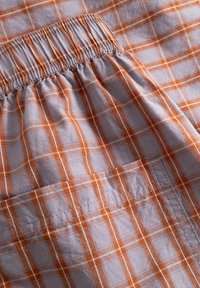 Ceinture élastique et poche rectangulaire sur un tissu à carreaux orange et blanc, montrant la couture détaillée et la texture du tissu.