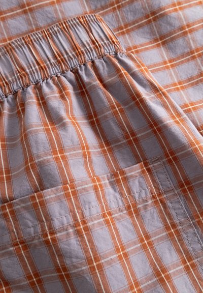 Ceinture élastique et poche rectangulaire sur un tissu à carreaux orange et blanc, montrant la couture détaillée et la texture du tissu.