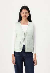 Ljusgrön gingham-väst med knäppning fram, två framfickor och strukturerad tyg. Buren över en vit långärmad topp.