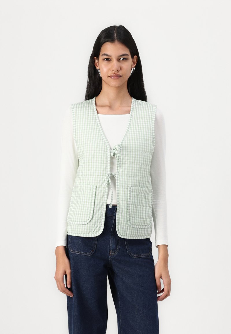 Ljusgrön gingham-väst med knäppning fram, två framfickor och strukturerad tyg. Buren över en vit långärmad topp.