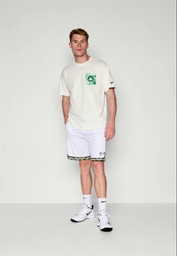 Mladic u bijelim Nike sportskim šortsevima, bijelom majici s zelenim logotipom, bijelim čarapama i bijelim Nike tenisicama stoji nasuprot jednostavne pozadine.