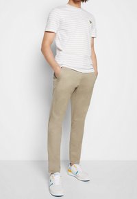 Camiseta de rayas gris claro con un pequeño logo, pantalones beige de corte slim y zapatillas blancas con acentos azules y amarillos. Conjunto casual.