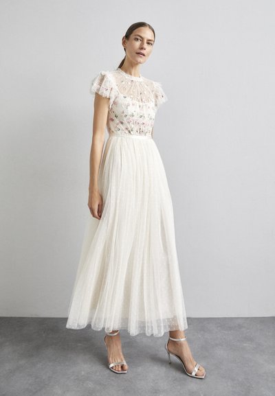 Witte, lange jurk met bloemenborduursel, doorschijnende, gerimpelde korte mouwen en een zwierige rok met stippen. Zilveren sandaaltjes met straps.