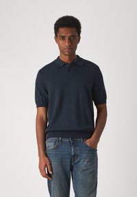 Polo shirt bleu marine en tissu tricoté avec un ourlet côtelé et des manches courtes, arborant un logo subtil sur la poitrine gauche. Porté avec un jean bleu.