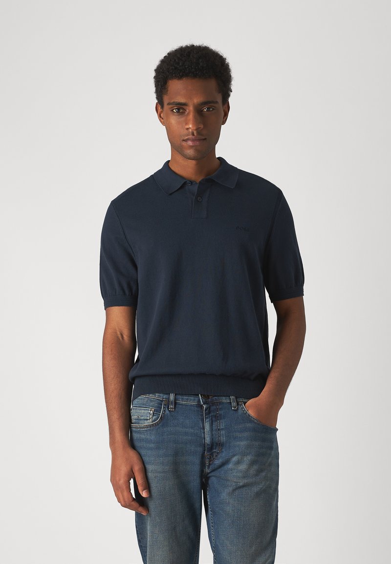 Polo shirt bleu marine en tissu tricoté avec un ourlet côtelé et des manches courtes, arborant un logo subtil sur la poitrine gauche. Porté avec un jean bleu.