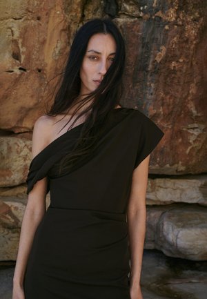 Femme aux longs cheveux noirs portant une robe noire à une épaule, debout devant un mur de roche texturé, les cheveux couvrant partiellement son visage.