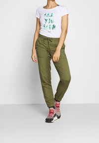 Vit t-shirt med grön bladtryck och texten "SEA YOU SOON", tillsammans med olivgröna joggerbyxor och flerfärgade vandringsskor.