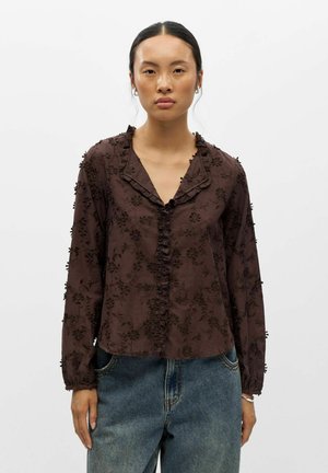 Femme aux cheveux foncés attachés, portant un chemisier marron à motifs floraux texturés à manches longues et un jean bleu ample, debout devant un fond uni.