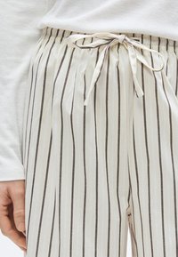 Pantalon de détente rayé en crème clair avec des rayures verticales marron foncé. Dispose d'une ceinture élastique avec un cordon de serrage noué et un tissu doux.