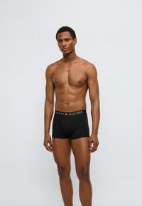 Μαύρα ανδρικά εσώρουχα μποξέρ με λογότυπο στη μέση, διακοσμημένα με κόκκινες και λευκές λεπτομέρειες, κατασκευασμένα από ελαστικό βαμβακερό ύφασμα. Το μοντέλο στέκεται όρθιο.