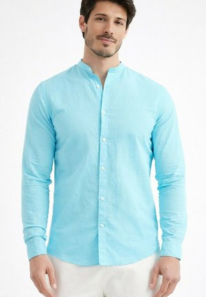 Homme portant une chemise à col mandarin bleu clair boutonnée, debout devant un fond blanc, faisant face à l'avant avec un léger sourire.