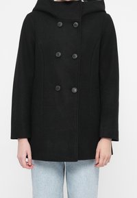 Black double-breasted coat with hood worn over light blue denim jeans, shown from shoulders to hips against a plain background.