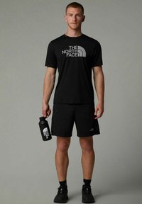 Camiseta negra y pantalones cortos con el logo de "THE NORTH FACE" en gris. El modelo sostiene una botella de agua negra y lleva zapatillas deportivas negras.