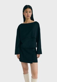Schwarzes cropped Oberteil mit langen Ärmeln und einem design mit herabhängenden Schultern, kombiniert mit einem schwarzen asymmetrischen Rock mit Knopfdetails.