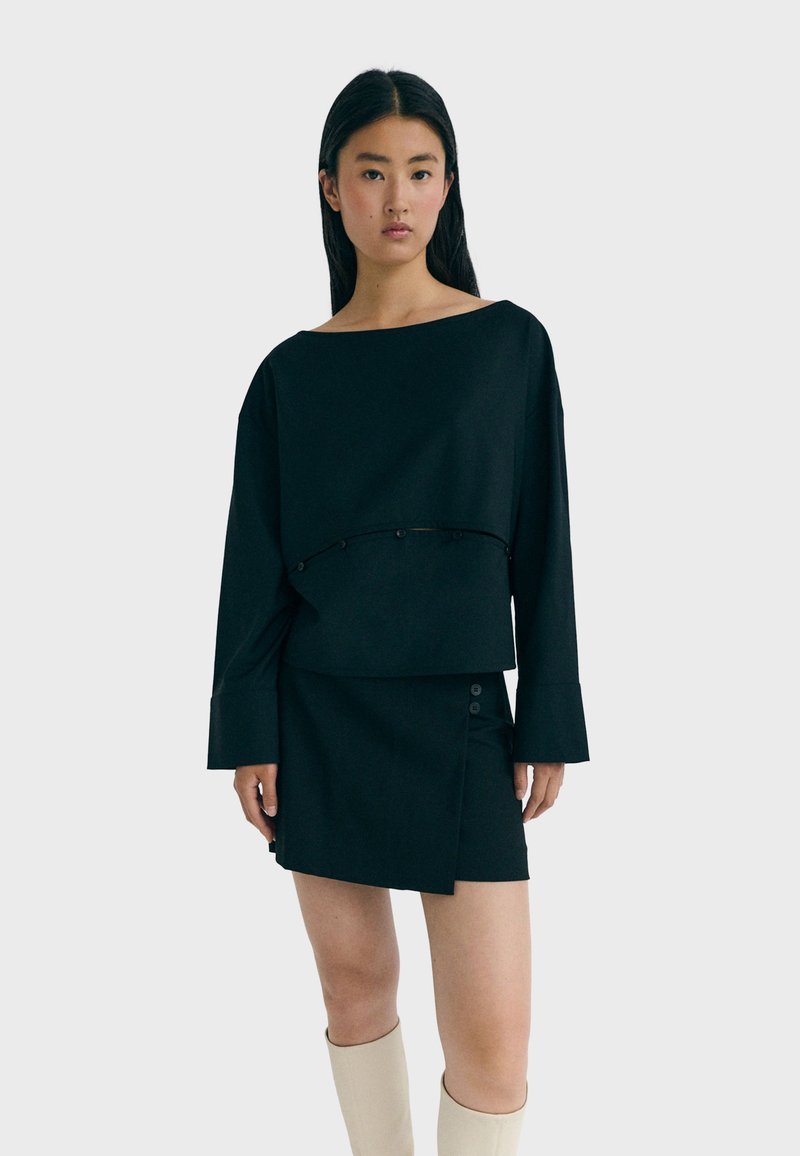 Schwarzes cropped Oberteil mit langen Ärmeln und einem design mit herabhängenden Schultern, kombiniert mit einem schwarzen asymmetrischen Rock mit Knopfdetails.