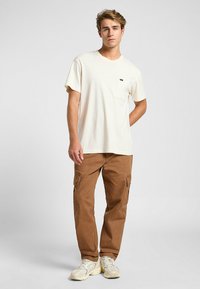 Camiseta crema de manga corta con bolsillo en el pecho, combinada con pantalones cargo marrones con bolsillos laterales. El modelo lleva zapatillas deportivas de color claro.