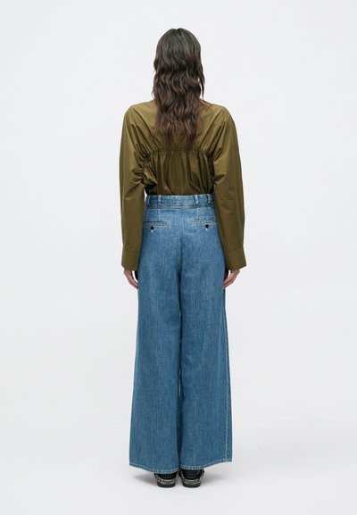 Πουκάμισο λαδί πράσινο, με πιέτες και μακριά μανίκια σε συνδυασμό με φαρδιά, ψηλόμεσα μπλε τζην παντελόνια. Μινιμαλιστικός σχεδιασμός χωρίς ορατά μοτίβα.