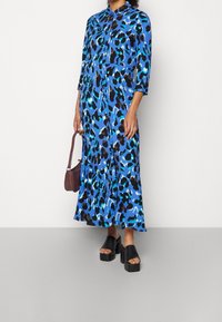 Maxikleid in Blau mit schwarzem und petrolfarbenem Leopardenmuster, vorne mit Knöpfen, lange Ärmel, fällt bis zur Mitte der Wade. Kombiniert mit schwarzen Sandalen mit klobigem Absatz.