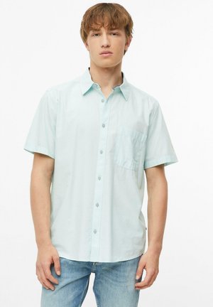 Jeune homme portant une chemise bleu clair à manches courtes boutonnée et un jean bleu, debout avec une expression neutre sur fond blanc.