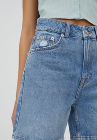 Pantalones cortos de mezclilla en azul claro con un acabado ligeramente descolorido. Presentan cierre de botón y cremallera, bolsillos delanteros y detalles desgastados.