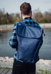 Dunkelblauer Rucksack mit einer glatten, matten Oberflächenstruktur und einem Klappenverschluss. Verfügt über gepolsterte Riemen und ein Logotag. Wird von einer Person in einem karierten Hemd getragen.