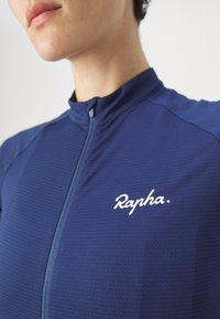 Gros plan d'une personne portant un haut zippé texturé bleu avec "Rapha" écrit en blanc sur la poitrine gauche.