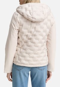 Veste matelassée beige clair avec un design matelassé et une capuche. Elle présente des manches lisses, un ourlet ajustable et un détail de couture subtil.