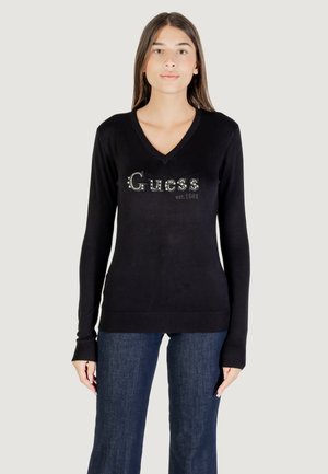 Black knit sweater with a V-neck and long sleeves. Features silver "Guess" logo with rhinestones. Ribbed hem and cuffs. Pair with blue jeans.