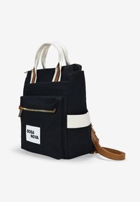 Mochila negra de lona con asas y correas en crema, bolsillos con cremallera y una etiqueta blanca con el texto "BOSA NOVA". Acentos de cuero marrón visibles.