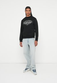 Čierna mikina s potlačou "JACK&JONES Coll. Studio" v bielej farbe, spárovaná so svetlomodrými džínami a bielymi teniskami s modrými akcentmi.