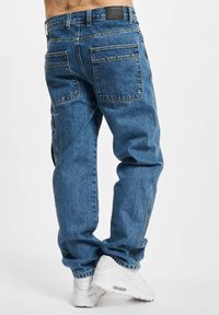 Denimjeans in Blau. Lockere Passform mit sichtbaren Nähten, zwei Gesäßtaschen und einem Etikett am Bund. Getragen mit weißen Sneakers.
