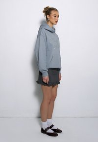 Jasnoniebieska bluza z kapturem z kieszenią na przodzie i haftowanym napisem, zestawiona z czarną plisowaną spódnicą z falbaną na dole oraz brązowymi butami.