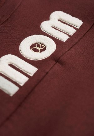 Bordeaux-Stoff mit weißem, strukturiertem gesticktem Schriftzug "Mama" und einem kleinen Logo-Akzent in Beige. Nahaufnahme mit Fokus auf Textur und Details.