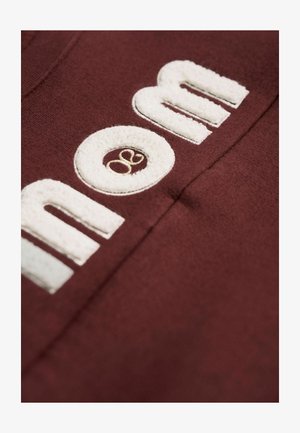 Tissu bourgogne avec des lettres blanches brodées texturées disant "maman" et un petit logo en beige. Gros plan sur la texture et les détails.