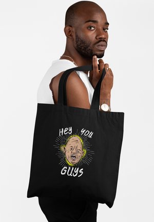 Homme barbu avec montre tenant un sac cabas noir avec un visage illustré et le texte « Hey you Guys » sur fond blanc.