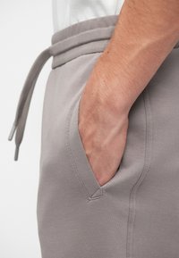 Pantaloni sport gri cu talie ajustabilă cu șnur și buzunar lateral, având o textură netedă și un design minimalist. Prim-plan al unei mâini în buzunar.