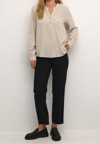 Chemise beige à manches longues avec un motif rayé, col henley, trois boutons, associée à un pantalon noir à jambes droites et des mocassins noirs.