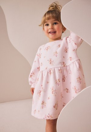 Usměvavá batole dívka se světle hnědými vlasy v drdolu, oblečená v růžových květinových šatech s dlouhým rukávem, stojící uvnitř poblíž zakřivených bílých tvarů.