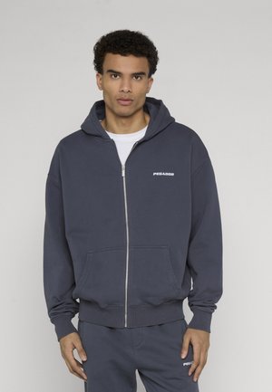 Jonge man die een marineblauwe hoodie met rits en bijpassende broek draagt met "PEGADOR"-logo's, staand tegen een effen lichte achtergrond.