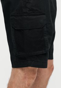 Short cargo noir avec poche à rabat recouverte et panneau en mesh sur le côté, porté sur la cuisse d’une personne.