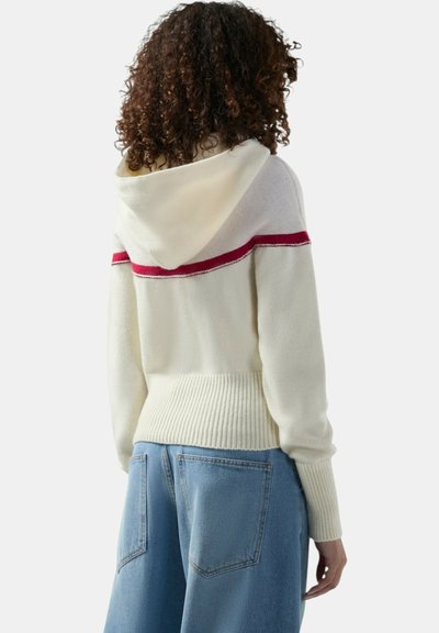 Persona con capelli ricci che indossa una felpa con cappuccio bianca a maglia con una striscia rossa sulle spalle e jeans a vita alta e gamba larga di colore azzurro chiaro, vista di spalle.