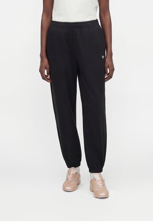 Personne portant un pantalon de jogging noir à taille élastique avec un petit logo sur la cuisse, des chaussettes blanches, des baskets rose clair et un top blanc sans manches.