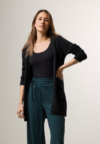 Svart cardigan med långa ärmar, framfickor, kombinerad med mörkgröna byxor med elastisk midja. Släta texturer, avslappnad design.