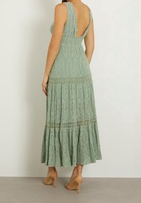 Mintgrön spetsmaxiklänning med låg rygg, breda axelremmar och lager på lager-skirt. Accenterad med intrikata spetsdetaljer och en flödande silhuett.