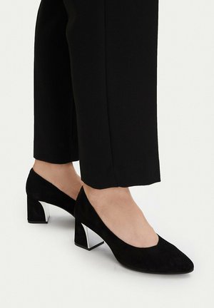 Nohy v černých semišových lodičkách na blokovém podpatku s kovovými doplňky, v kombinaci s černými rovnými kalhotami ke kotníkům na bílém pozadí.