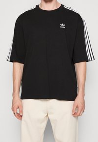 Svart bomullströja med vit tre-stra stripe-design på ärmarna och liten Adidas-logotyp på bröstet. Avslappnad passform, rund halsringning, korta ärmar.